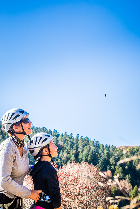 Adulte et enfant équipés de casques observant une personne en tyrolienne dans un paysage de montagne.