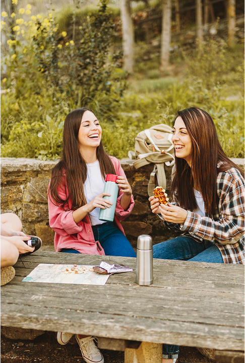 Deux jeunes femmes randonneuses riant autour d’un goûter en pleine nature, assises à une table en bois.