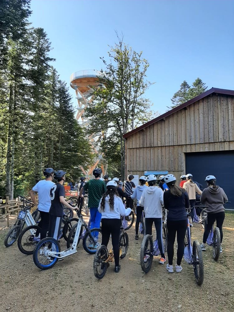 Groupe de personnes équipées de casques et de trottinettes tout-terrain, rassemblées près d’un bâtiment en bois au parc de Prabouré.