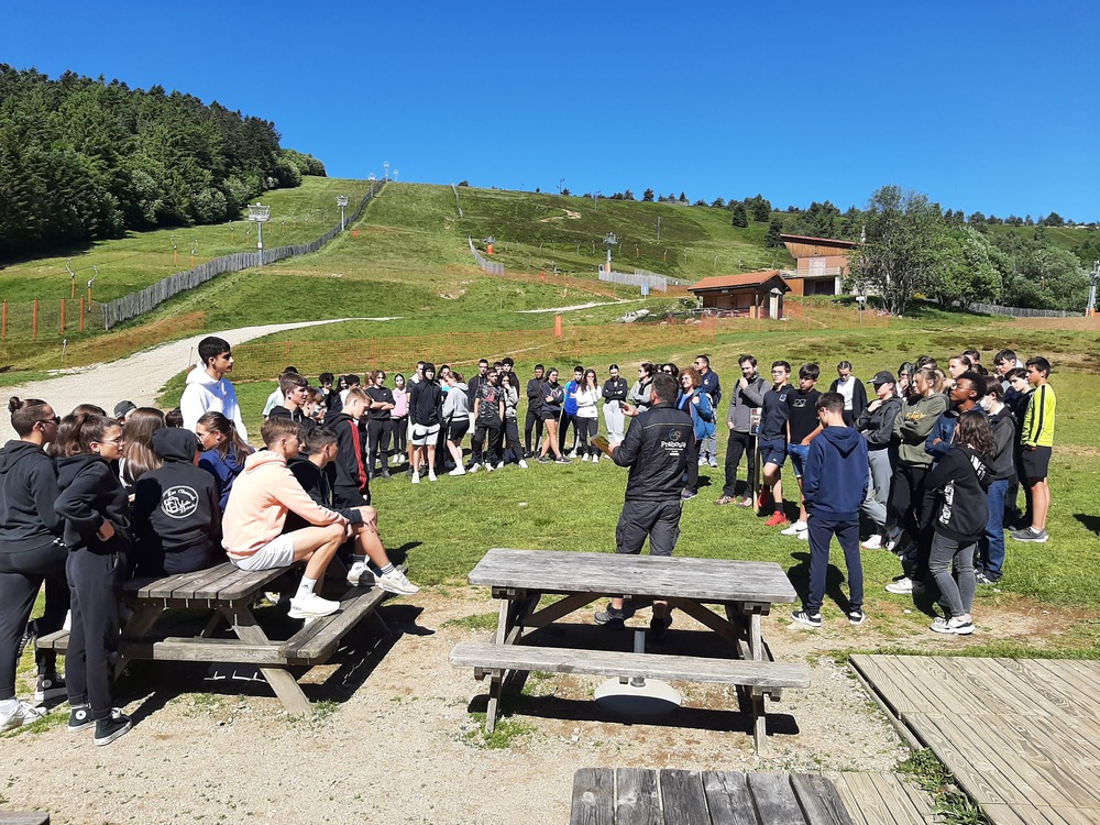 Grand groupe de jeunes rassemblés en plein air autour d’un animateur au parc de Prabouré, avec les pistes et remontées mécaniques visibles à l’arrière-plan.