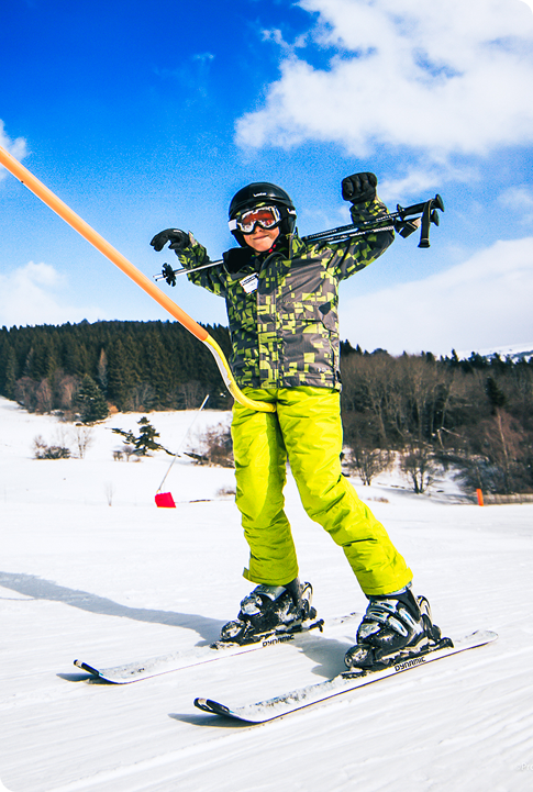 Skieur portant un casque et une tenue verte fluo tenant ses bâtons en l’air, souriant sur une piste enneigée avec des sapins en arrière-plan.