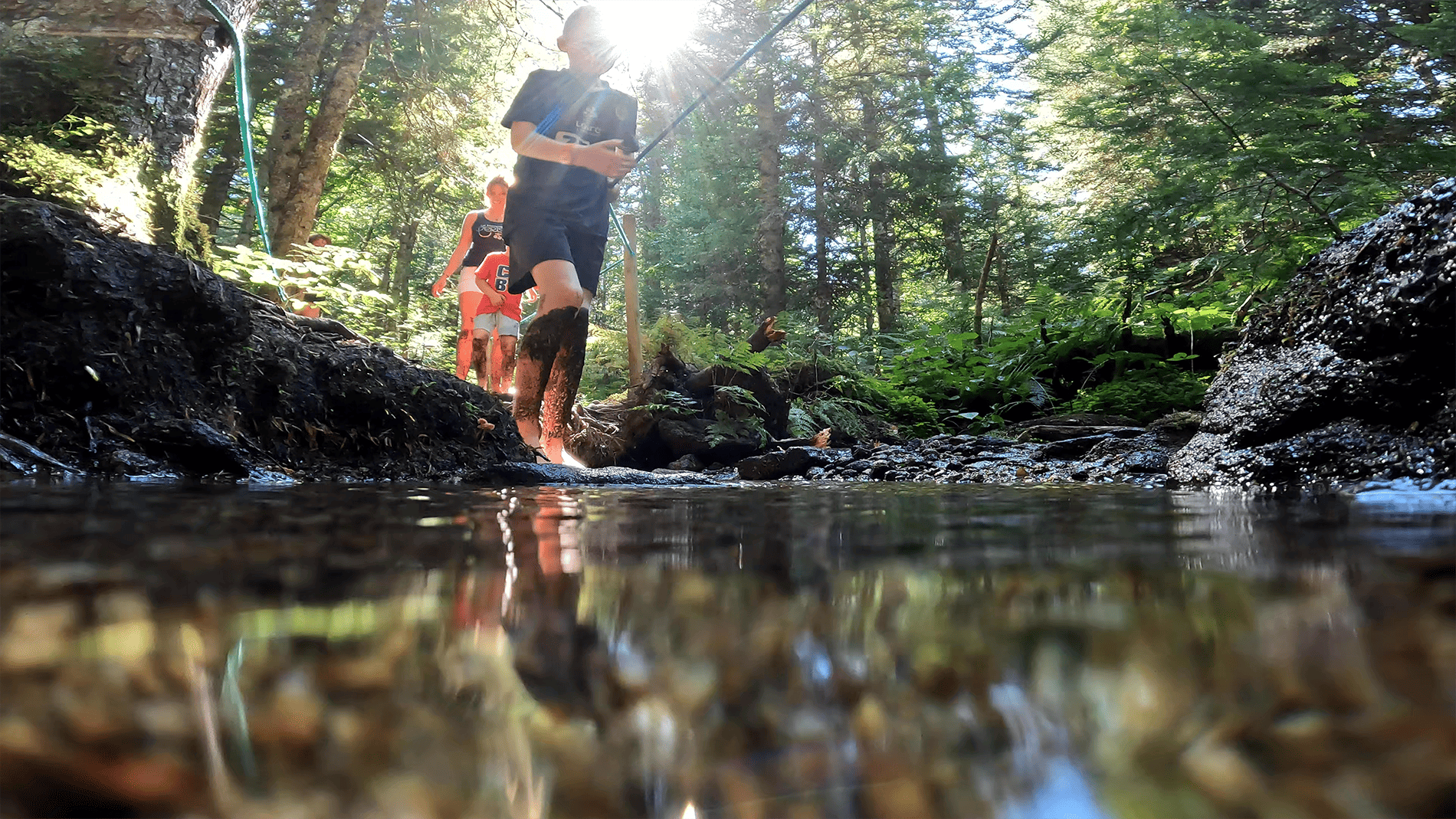 Deux enfants et une femme marchant pieds nus dans un ruisseau en forêt, les jambes couvertes de boue, sous un rayon de soleil filtrant à travers les arbres.