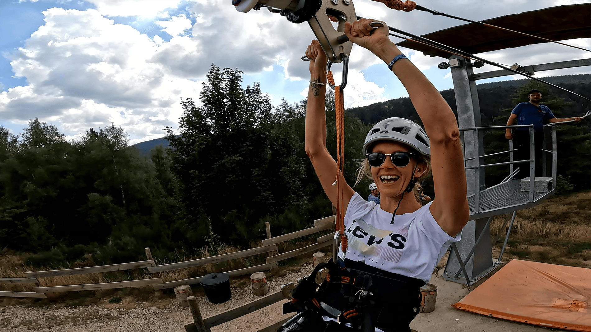 Femme souriante portant un casque et des lunettes de soleil, suspendue à une tyrolienne à l’arrivée d’une plateforme, par temps partiellement nuageux.