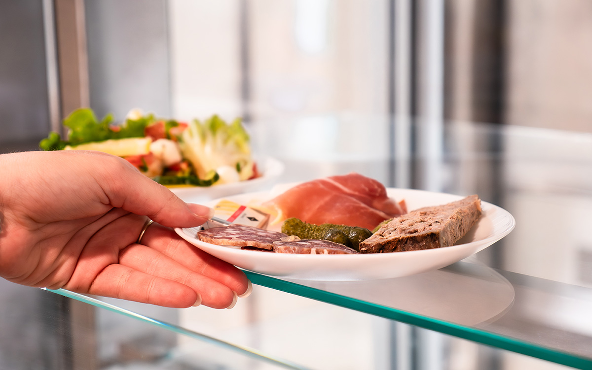 Main prenant une assiette de charcuterie et cornichons dans une vitrine réfrigérée, avec un autre plat de salade en arrière-plan.
