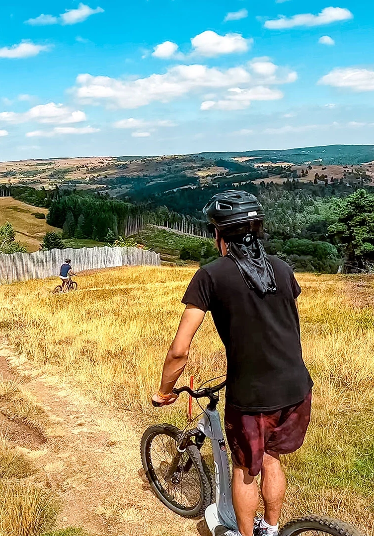 Personne en tenue de sport et casque descendant un sentier à trottinette tout-terrain à Prabouré, offrant une vue panoramique sur les collines et forêts environnantes.