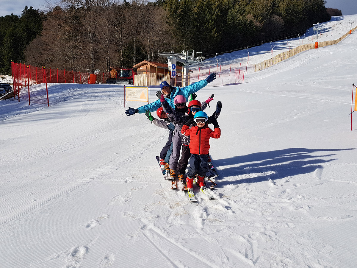 Groupe d’enfants en combinaison de ski et casque, alignés sur la neige et levant les bras en souriant, sur une piste ensoleillée bordée d’arbres au parc de Prabouré.
